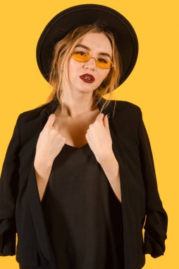 Siyah ceketli, güneş gözlüklü ve sarı arka planda şapkalı beyaz bir kadın.