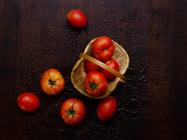Tomatoes in a wicker basket and scattered on a dark wooden surface, with water drops, close-up, top view