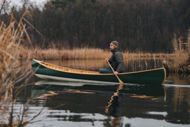Man paddling a beautiful green canoe on the pond, calm late autumn nature. Active lifestyle, a person enjoying tranquil early spring scenery on the water