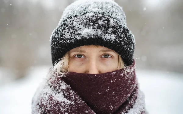 Blonde woman in beanie hat and nose covered in scarf looks at camera in winter snowfall. Nordic winter seasonal portrait, tranquil and calm outdoor scene