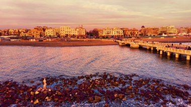 Sunset Rome beach aerial view from sea over cliff and ruin of Neptune statue with city skyline with downtown with Ravennati square and pedestrian pier a landmark of tourist and residents