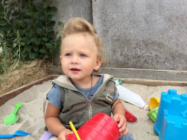 Kum havuzunun yanında fotoğrafçı pozu veren güzel bir bebek. Bebek kum havuzundaki kuru kum tanelerinden oluşur. Küçük çocuklar için ev yapımı kum havuzu. Kumlu bebek kum havuzu, kum havuzu ve çocuk oyunu..