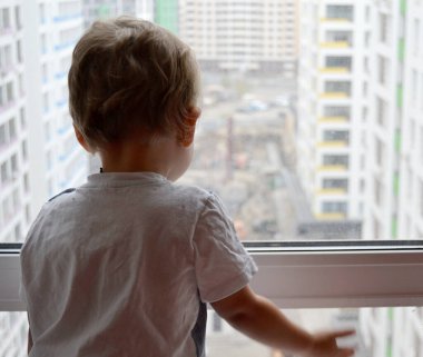 Renkli fotoğraf için fotoğrafçı pozu veren çocuk yüzlü güzel bir erkek bebek. Cadre, pencerenin yanında beyaz bir ifadeyle çocuk görünümünde bir çocuktan oluşuyor. Sağlıklı erkek bebek. Pencerenin yanındaki çocuk.
