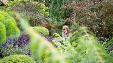 Güzel genç bir kadın, yemyeşil çiçeklerle çevrili bir bahçede taş basamaklarda oturuyor. Düşünceli dinlenen kız.