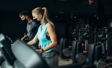 Modern spor salonunda koşu bandında koşan genç, zinde bir kadın ve erkek. Mesafelerini koruyorlar ve koruyucu yüz maskeleri takıyorlar. Coronavirus dünya salgını ve spor teması.