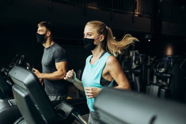 Modern spor salonunda koşu bandında koşan genç, zinde bir kadın ve erkek. Mesafelerini koruyorlar ve koruyucu yüz maskeleri takıyorlar. Coronavirus dünya salgını ve spor teması.
