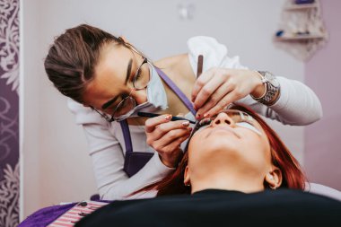 Kirpik uzatma prosedüründeki güzel kızıl saçlı kadın. Kozmetik ve vücut bakımı yakın çekim. Koruyucu yüz maskesi takan kadın güzellik uzmanı. Seçici odak.