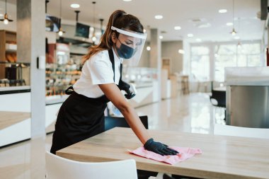 Fırında ya da fast food restoranında çalışan güzel bir kadın. Coronavirüs salgınına karşı masaları temizleyip dezenfekte ediyor. Koruyucu maske, eldiven ve yüz maskesi takıyor..