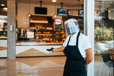 Beautiful woman with face shield and protective mask working bakery or fast food restaurant. She is standing in front shop doors and welcoming new customers. Coronavirus or Covid-19 safety measures. clipart