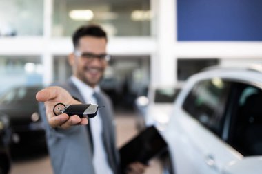 Yakışıklı, neşeli ve arkadaş canlısı bir satıcı araba kuaföründe veya galeride poz verir. Kameraya bakarken arabanın anahtarlarını tutuyor. Eldeki seçici odaklanma.