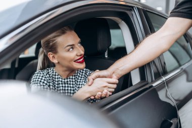 Satisfied and smiled female buyer sitting in her new car. She is smiling, looking at seller through open window while handshaking with him.