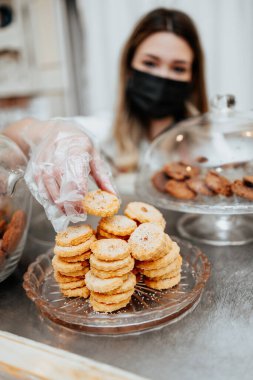Koruyucu maskeli güzel bir bayan satıcı bir şekerleme dükkanında ya da fırında çalışıyor. Lezzetli tatlılar satıyor ve paketliyor. Coronavirus ve küçük işletme konsepti.