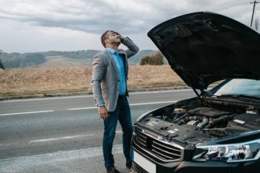 Orta yaşlı şık bir işadamı araba arızasını veya motor arızasını düzeltmeye çalışıyor ve yolda araba kazası için çekici servisi bekliyor. Yol kenarı yardım kavramı.