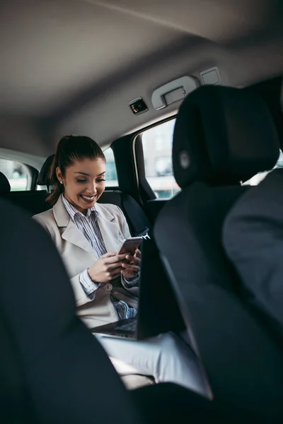 漂亮的年轻女商人坐在豪华轿车后座上 她用她的智能手机和微笑 公司商业概念中的运输 — 图库照片