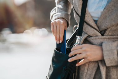 Sıradan giyinmiş bir iş kadınının, akıllı telefonunu çantasından çıkartırken çekilmiş yakın plan fotoğrafı. Parlak güneşli bir gün ve arka planda güzel bir bokeh..