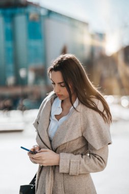 Sıradan giyinmiş bir iş kadını şehirde dikiliyor ve akıllı telefonunu biriyle iletişim kurmak için kullanıyor. Parlak güneşli bir gün ve arka planda güzel bir bokeh..