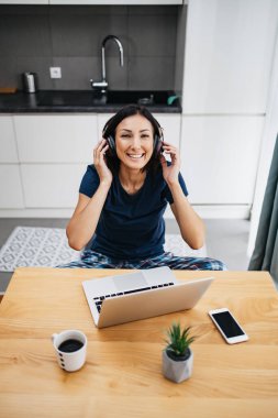 Çekici ve mutlu orta yaş kadın serbest çalışanı evinde çalışıyor. O kulaklık kullanıyor, müzik dinliyor ve şarkı söylüyor. Arka planda modern mutfak. Serbest meslek kavramı.