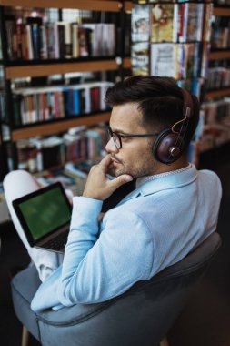 Orta yaşlı yakışıklı adam modern kitapçıda oturuyor, kulaklıkla müzik dinlerken dizüstü bilgisayarını ya da bilgisayarını okuyor ve kullanıyor..