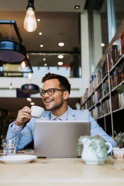 Genç yakışıklı adam modern kitapçıda oturuyor, dizüstü bilgisayarını veya bilgisayarını okuyor ve kullanıyor..