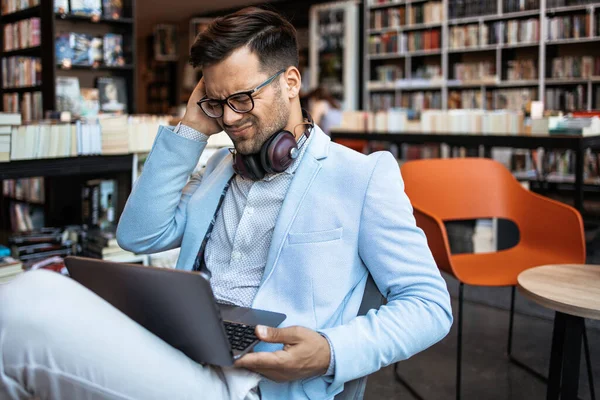 Orta yaşlı yakışıklı adam modern kitapçıda oturuyor, kulaklıkla müzik dinlerken dizüstü bilgisayarını ya da bilgisayarını okuyor ve kullanıyor..