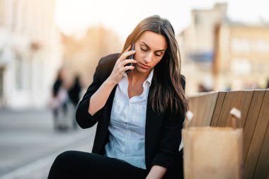 Kendine güvenen genç iş kadını sokak bankında oturuyor ve bilgisayarını iş ve iletişim için kullanıyor. O ciddi ve yana bakıyor. İş ve mobil ofis kavramı. Parlak güneşli bir gün. 