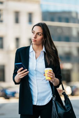 Sıradan giyinmiş bir iş kadını şehrin caddesinde duruyor ve elinde kahve tutarken biriyle iletişim kurmak için akıllı telefonunu kullanıyor. Parlak güneşli bir gün.