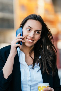 Sıradan giyinmiş bir iş kadını şehrin caddesinde duruyor ve elinde kahve tutarken biriyle iletişim kurmak için akıllı telefonunu kullanıyor. Parlak güneşli bir gün.