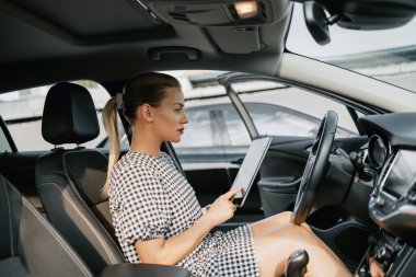 Beautiful young woman working as used car seller. She is using digital tablet and checking car condition before customer or buyer. Used vehicle dealership.
