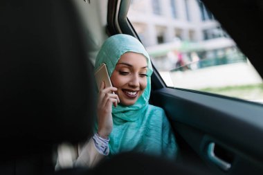 Lüks arabada arka koltukta oturan başörtüsü ile güzel Müslüman Arap kadın. Akıllı telefonunu kullanıyor ve pencereden bakıyor..