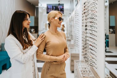 Modern optik mağazada gözlük çerçevesi seçen güzel ve modaya uygun bir kadın. Kadın satıcı uzmanı doğru karar vermesine yardımcı oluyor..