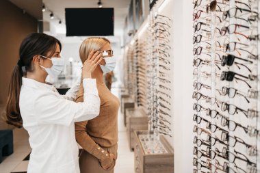 Modern optik mağazasında gözlük çerçevesi seçen sarışın bir kadın. Kadın satıcı uzmanı doğru karar vermesine yardımcı oluyor. Virüs salgınına karşı koruyucu yüz maskesi takıyorlar..