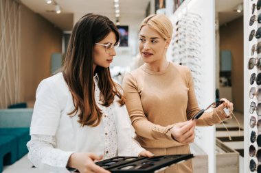 Modern optik mağazada gözlük çerçevesi seçen güzel ve modaya uygun bir kadın. Kadın satıcı uzmanı doğru karar vermesine yardımcı oluyor..