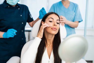 Güzel ve mutlu genç bir kadın tıbbi sandalyede oturup aynaya bakıyor. Hyaluronik asit doldurucu ya da botulinyum toksin iğneleriyle başarılı bir güzellik tedavisinden sonra tatmin oluyor..