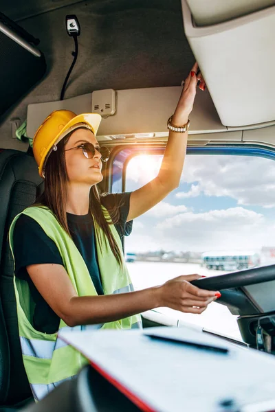 Portrait of beautiful young woman professional truck driver with protective yellow helmet sitting and driving a big truck. Inside of vehicle. People and industrial transportation concept.