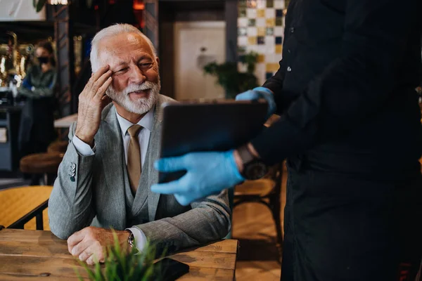 The waiter taking order for a delicious meal to the senior businessman at the restaurant. He wears a protective mask as part of security measures against the Coronavirus pandemic.