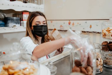 Koruyucu maskeli güzel bir bayan satıcı bir şekerleme dükkanında ya da fırında çalışıyor. Lezzetli tatlılar satıyor ve paketliyor. Coronavirus ve küçük işletme konsepti.