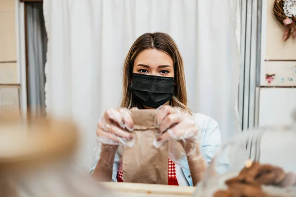 Beautiful young female seller with protective face mask working in a confectionery shop or bakery. She is selling and packing delicious sweets. Coronavirus and small business concept.