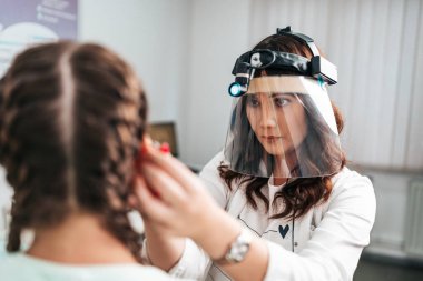 Audiologist doing impedance audiometry or diagnosis of hearing impairment. An beautiful teenage girl getting an auditory test at a hearing clinic. Doctor wearing face shield against Covid-19 virus.