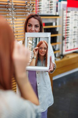 Beautiful and fashionable redhead woman choosing eyeglasses frame in modern optical store. Female seller specialist helps holding a mirror and helping her to make right decision.