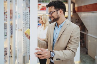 Genç bir iş adamı eczaneden ya da eczaneden ilaç alırken çekici bir kadın eczacı arka planda bir şeyler yapıyor..