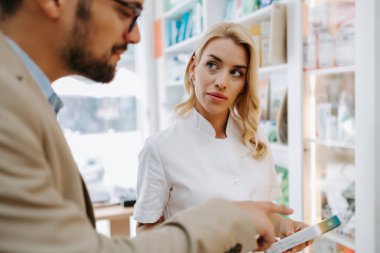 Young business man choosing and buying drugs in a drugstore while talking with attractive female pharmacist. She helping him with expert advice.