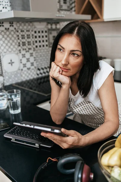 Unhappy and worried middle age woman sitting alone in her apartment and using laptop computer or tablet. She is bored because social distancing during pandemic quarantine.