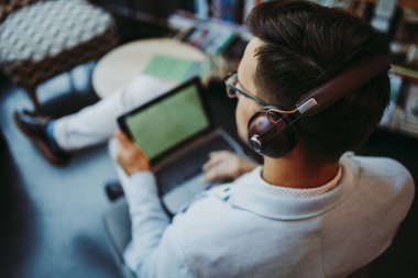 Orta yaşlı yakışıklı adam modern kitapçıda oturuyor, kulaklıkla müzik dinlerken dizüstü bilgisayarını ya da bilgisayarını okuyor ve kullanıyor..