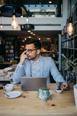 Genç yakışıklı adam modern kitapçıda oturuyor, dizüstü bilgisayarını veya bilgisayarını okuyor ve kullanıyor..