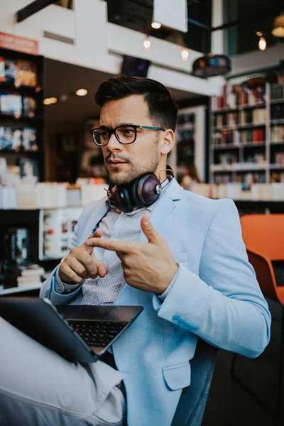 Orta yaşlı yakışıklı adam modern kitapçıda oturuyor, kulaklıkla müzik dinlerken dizüstü bilgisayarını ya da bilgisayarını okuyor ve kullanıyor..