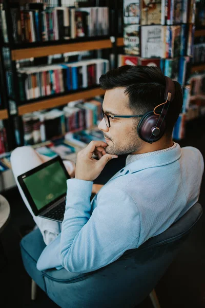 Orta yaşlı yakışıklı adam modern kitapçıda oturuyor, kulaklıkla müzik dinlerken dizüstü bilgisayarını ya da bilgisayarını okuyor ve kullanıyor..