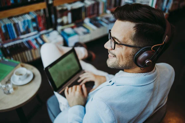 Orta yaşlı yakışıklı adam modern kitapçıda oturuyor, kulaklıkla müzik dinlerken dizüstü bilgisayarını ya da bilgisayarını okuyor ve kullanıyor..