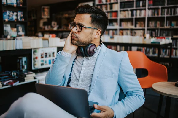 Orta yaşlı yakışıklı adam modern kitapçıda oturuyor, kulaklıkla müzik dinlerken dizüstü bilgisayarını ya da bilgisayarını okuyor ve kullanıyor..