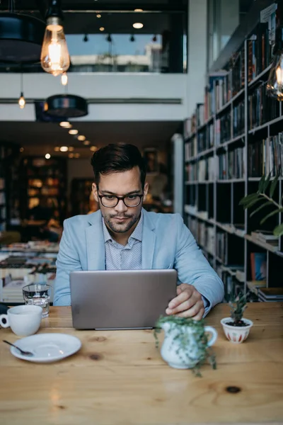 Genç yakışıklı adam modern kitapçıda oturuyor, dizüstü bilgisayarını veya bilgisayarını okuyor ve kullanıyor..
