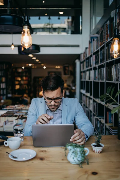 Genç yakışıklı adam modern kitapçıda oturuyor, dizüstü bilgisayarını veya bilgisayarını okuyor ve kullanıyor..